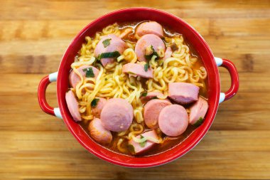 Ahşap arka planda kırmızı tabakta sosisli ramen eriştesi. Her zamanki ucuz yemekler. Ucuz ve lezzetli yemek kavramı.