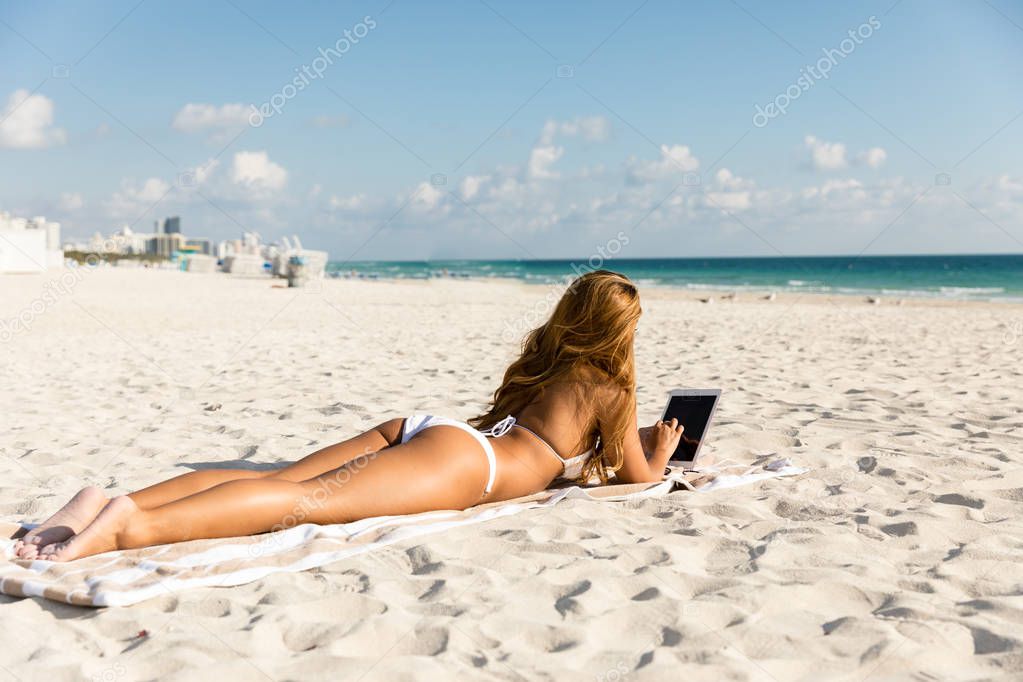 Attractive woman lying down on beach by the ocean using a tablet