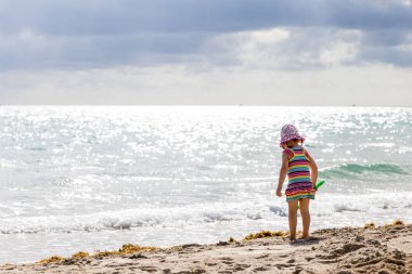 Küçük kız bir sundress ve şapka sahilde görünüyor 