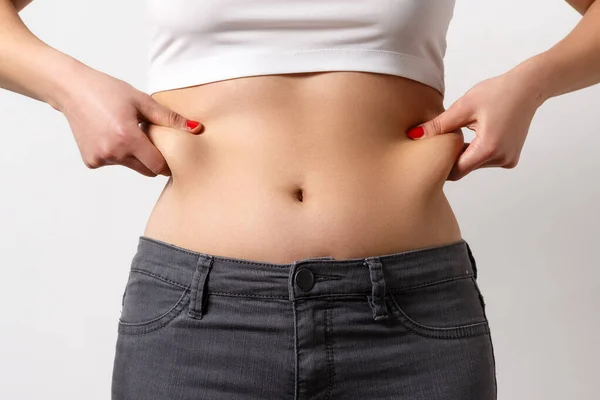 Kadın parmaklarıyla karnındaki ve yanlarındaki fazlalıkları çimdikliyor. Beyaz bir arkaplanda, yakın plan