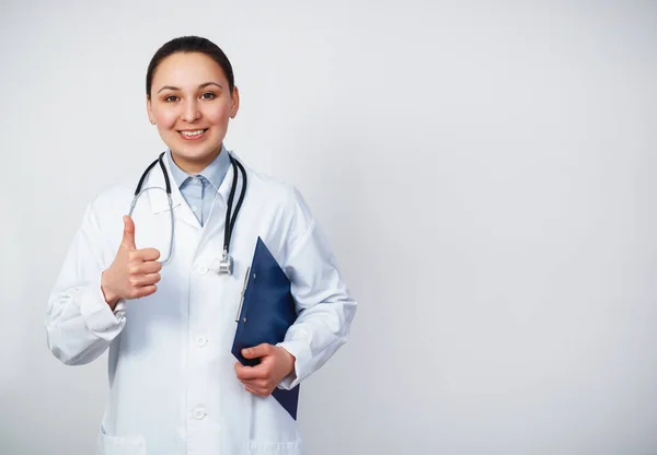 白い背景に孤立した親指を示す聴診器で笑顔の若い医師の女性 ノートパッドフォルダ上の健康カードを保持医療ガウンの女性医師 医療従事者の概念 — ストック写真
