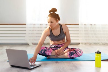 Genç tatlı kız evde yoga yapıyor, poz veriyor ve dizüstü bilgisayara bakıyor. Pencerenin dışında güneş parlıyor..