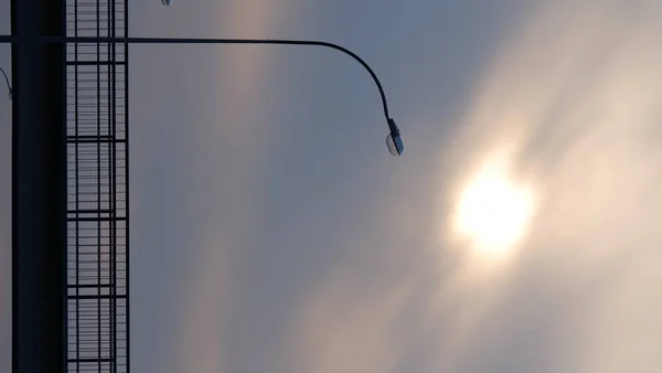 Lonely lantern on the background of the sun, shining through the cloudy sky. — Stock Photo, Image