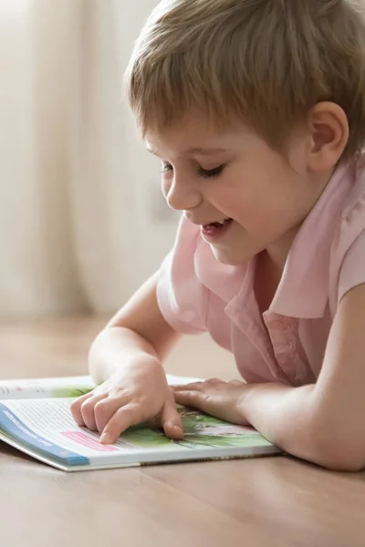 Un niño pequeño y lindo, tumbado en el cálido piso de su casa, leyendo una historia interesante en un libro enorme . — Foto de Stock