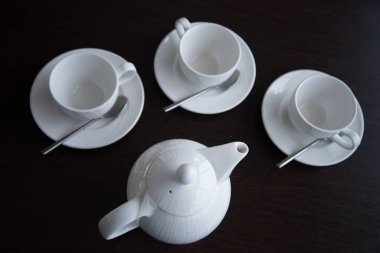 Hafif çay tabakları koyu bir masaya konur. Bir beyaz çaydanlık ve üç fincan çay tabağı ve kaşıkla..