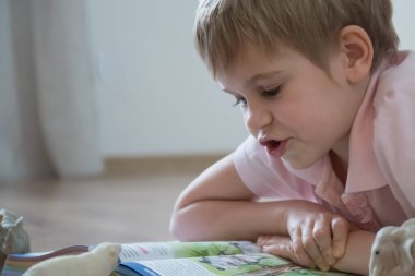 Kısa saçlı güzel bir sarışın çocuk büyük bir kitap okuyor. Anaokulu öğrencisi sıcak zemine oturdu. Konforlu ve mutlu ev ortamı.