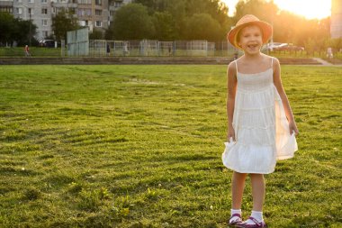 Mutlu ve gülümseyen, şapkalı güzel bir kızın güneşin batışındaki kameraya gülümseyen bir portresi. Şehirde yaz tatili. Yaz zamanı. Yaşam tarzı. Pozitif duygular