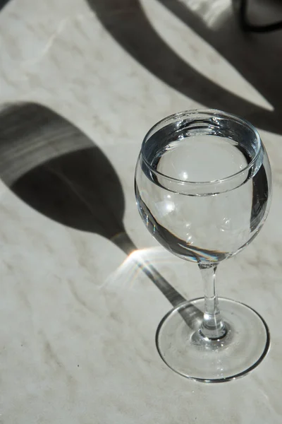 Ett högt glas står på bordet i solen. Glaset bildar en skugga, ett intressant geometriskt mönster. Det finns vatten i glaset. Vertikal inriktning — Stockfoto