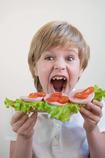 A child with his mouth wide open. Bites into a large ham and vegetable sandwich. School lunch. Portrait