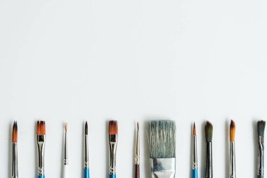 Altta, temiz beyaz bir çarşafın üzerinde, boyalı çizimler için fırçalar var. Farklı şekil ve boyutlarda fırçalar. Kartpostal, geçmiş, doku. Yukarıdan görünüşü.