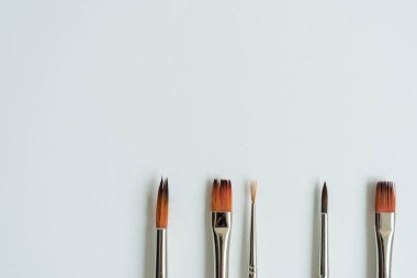 Altta, temiz beyaz bir çarşafın üzerinde, boyalı çizimler için fırçalar var. Farklı şekil ve boyutlarda fırçalar. Kartpostal, geçmiş, doku. Yukarıdan görünüşü.