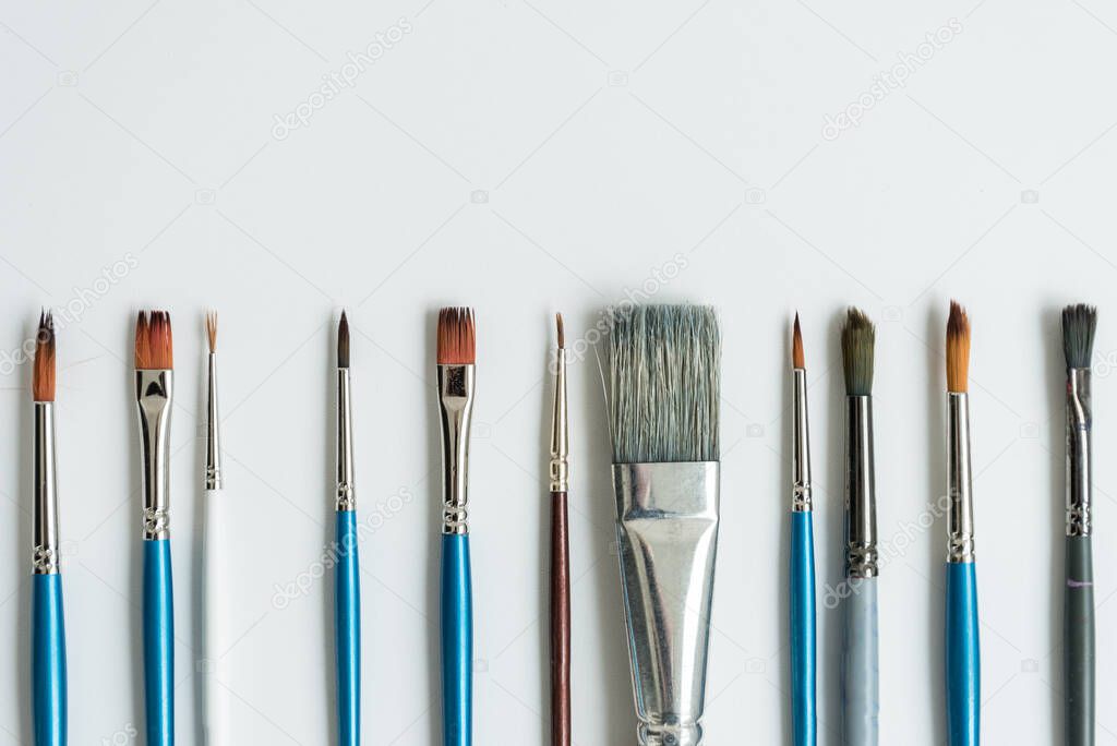 At the bottom, on a clean white sheet, there are brushes for drawing with paints. Brushes of different shapes and sizes. Postcard, background, texture. The view from the top.