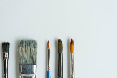 Altta, temiz beyaz bir çarşafın üzerinde, boyalı çizimler için fırçalar var. Farklı şekil ve boyutlarda fırçalar. Kartpostal, geçmiş, doku. Yukarıdan görünüşü.