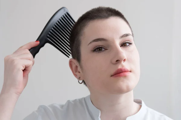 A young brunette girl with short hair is combing her hair with a huge comb. Bald.