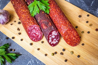 Tahta kesme tahtasının üzerinde üç çeşit lezzetli füme sosis. Yakınlarda biber, maydanoz ve sarımsak var. Koyu mermer arkaplan.