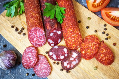 Tahta kesme tahtasının üzerinde üç çeşit lezzetli füme sosis. Yakınlarda biber, maydanoz, sarımsak ve domates var. Koyu mermer arkaplan.