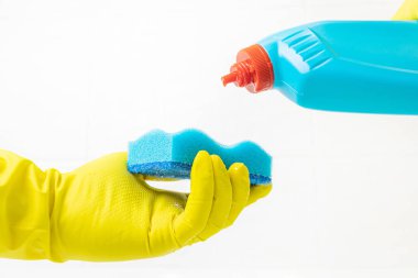 Mavi süngerli sarı lateks eldivenli kadın eli. Öte yandan bir banyo temizleyicisi. Arkaplan - beyaz fayans. Banyoyu temizleme konsepti, temizlik. Kopyalama alanı.