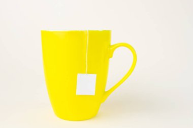Sarı fincanlı çay poşeti. Bardaktan beyaz bir etiket sarkıyor. Hafif pastel arkaplan.