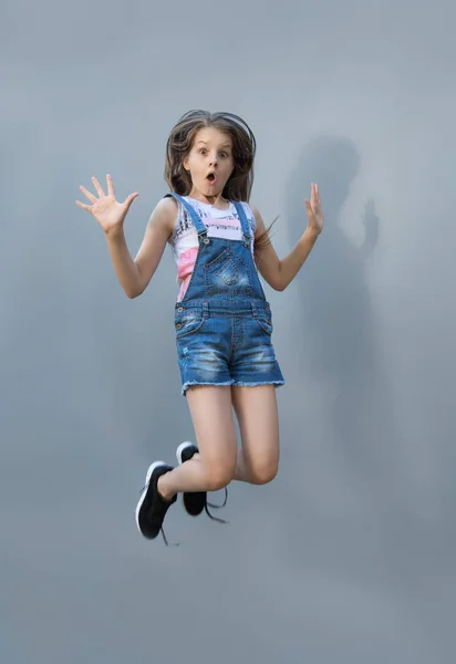Dziewczyna radośnie skoki. Wesoła Dziewczyna Nastolatka w Dżinsowe ogrodniczki na szarym tle — Zdjęcie stockowe