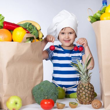 Büyük şef şapkalı küçük mutlu kız bir turp tutan ürünler çanta. Taze meyve ve sebze masada torbalarda çeşitli.