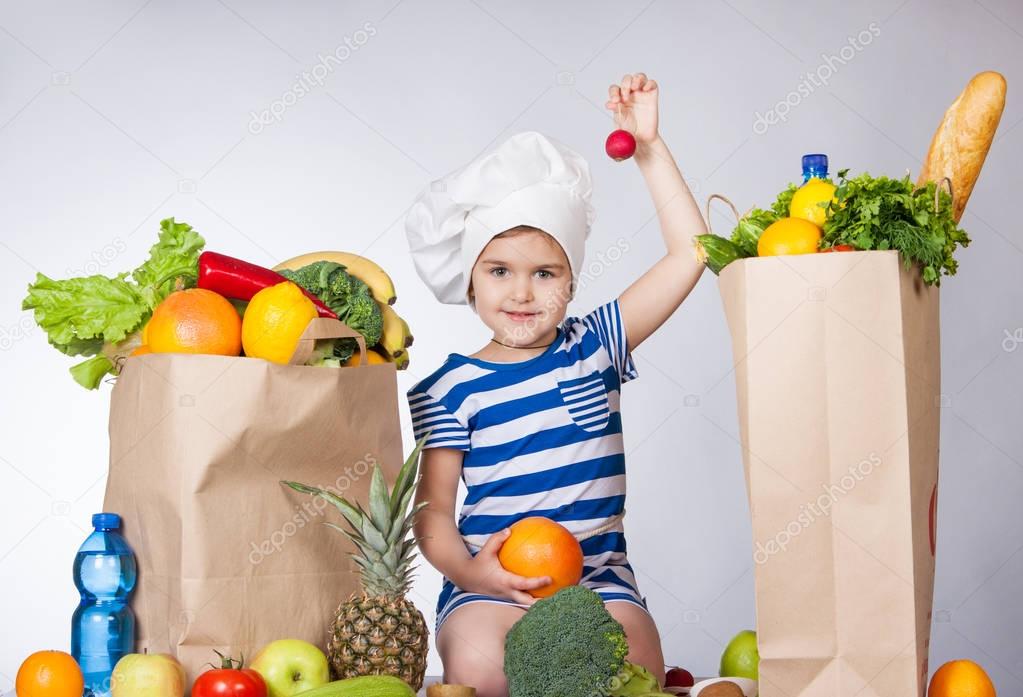 Little happy girl in the hat of the chef with big bags products holding a radish. A variety of fresh fruits and vegetables in bags on the table.