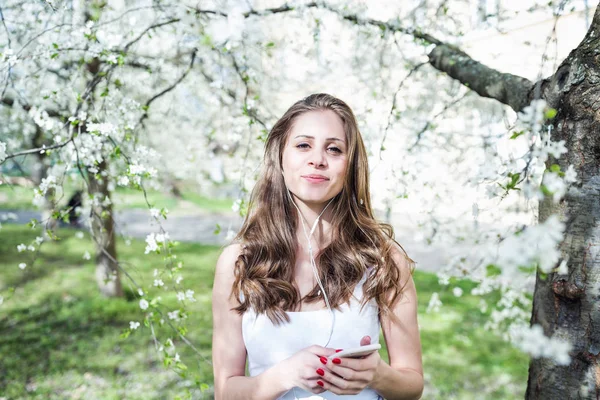 Junge Frau lächelt und hört Musik oder Podcast über Kopfhörer unter den blühenden Bäumen. — Stockfoto