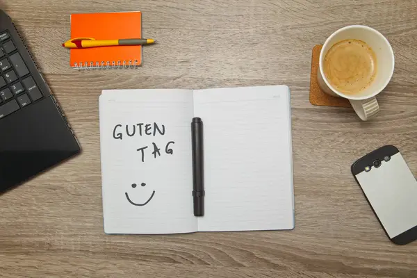 Cuaderno abierto con texto alemán "GUTEN TAG" (Buenas tardes) y una taza de café sobre fondo de madera. Vista de arriba hacia abajo — Foto de Stock