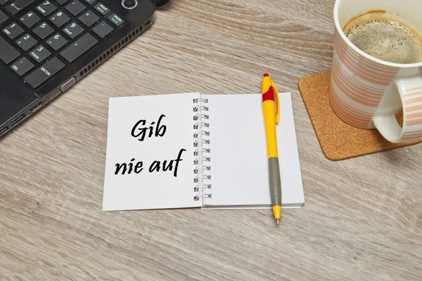 Cuaderno abierto con texto alemán "Gib Nie Auf" (Nunca te rindas) y una taza de café sobre fondo de madera. Vista de arriba hacia abajo — Foto de Stock