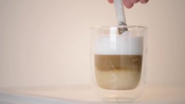 Yakın plan kadın eli, temiz cam bardakta kaşıkla taze macchiato kahvesi karıştırıyor..
