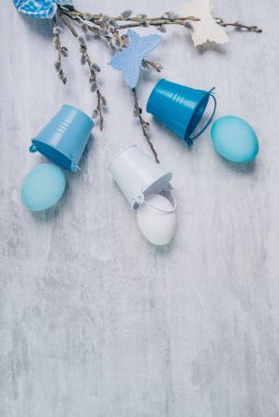 Mavi paskalya yumurtaları ve söğüt dalları mavi kurdeleyle bağlanmış ve gri ahşap bir masada küçük kovalarla. Üst görünüm, metin için boşluk.