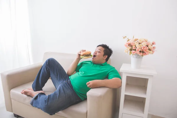 Líný obézní člověk jíst hamburger i když leží na gauči — Stock fotografie