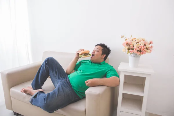 Perezoso hombre con sobrepeso comiendo hamburguesa mientras está acostado en un sofá —  Fotos de Stock