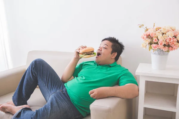 Preguiçoso homem com sobrepeso comer hambúrguer enquanto deitado em um sofá — Fotografia de Stock