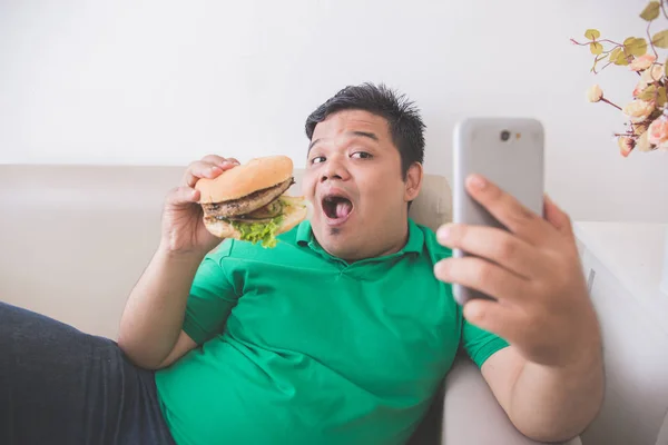 obese person eating hamburger while using mobile phone