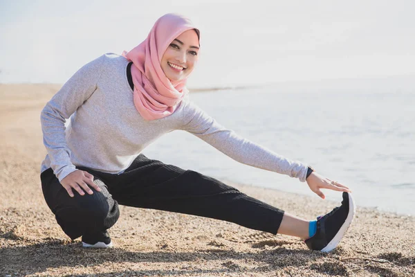 足のストレッチをしながら笑みを浮かべてヒジャーブを着ているアジアのスポーティな女性 — ストック写真