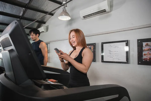 Jóvenes asiático muscular mujeres ejecutar en cinta de correr celebración de teléfono inteligente —  Fotos de Stock