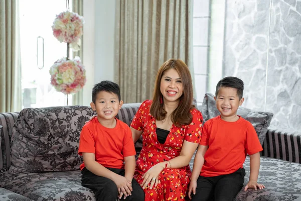 笑着坐在沙发上的亚裔母亲和两个穿着红色主题衣服的儿子 — 图库照片