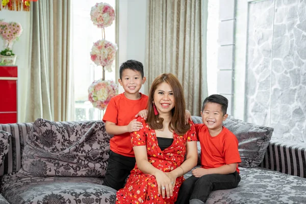 笑着坐在沙发上的亚裔母亲和两个穿着红色主题衣服的儿子 — 图库照片