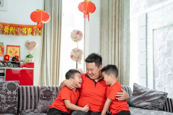 笑着坐在沙发上的中国父亲和两个穿着红色主题衣服的儿子 — 图库照片