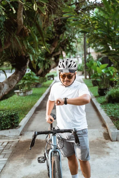Азиатский молодой человек в шлеме ходить с дорожным велосипедом, глядя на ее часы — стоковое фото