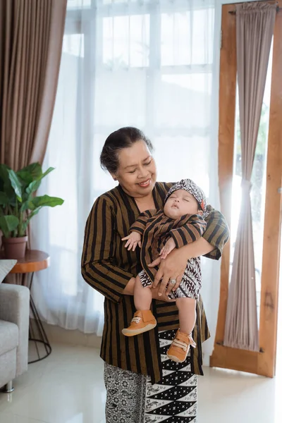 Nenek tersenyum asia membawa cucu kecil yang tertidur mengenakan batik javanese — Stok Foto