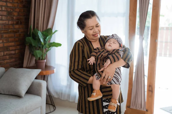 asian smiling grandmother carrying sleeping little grandson wearing javanese batik