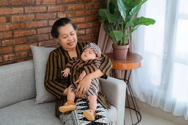 Nenek membawa cucu kecil yang tertidur ketika duduk di sofa, mengenakan batik jawa — Stok Foto