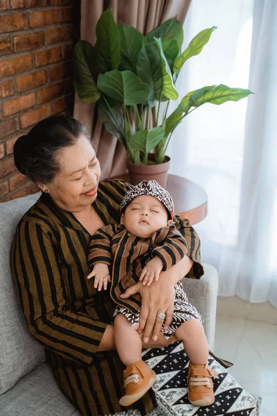 Azjatycki uśmiech babcia niosąc śpiący mały wnuk noszenie javanese batik — Zdjęcie stockowe