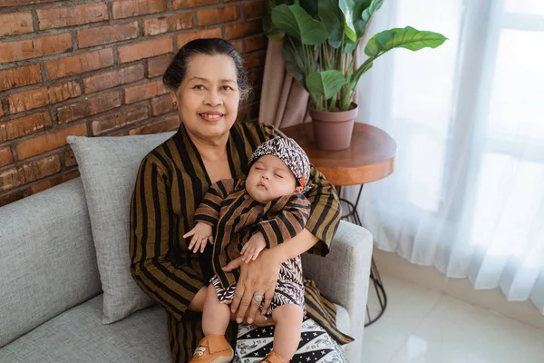 Nenek membawa cucu kecil yang tertidur ketika duduk di sofa, mengenakan batik jawa — Stok Foto