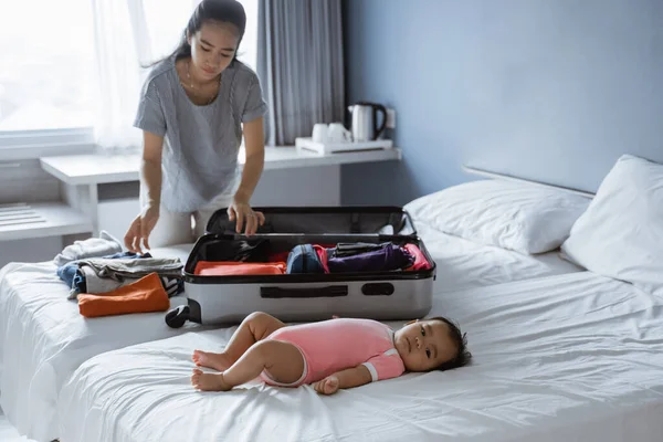 母親がスーツケースに身を包んだとき赤ん坊はベッドの上に横たわっています — ストック写真