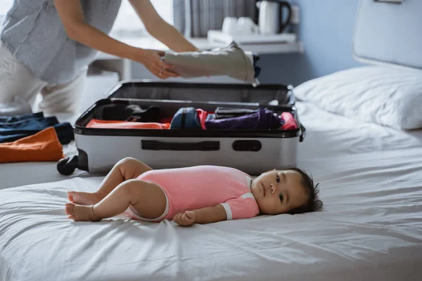 Bebê bonito deitado na cama ao lado de uma mala cheia — Fotografia de Stock