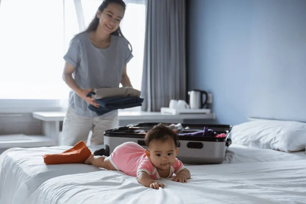 Lindo bebé se encuentra en la cama cuando su madre pone su ropa en una maleta — Foto de Stock