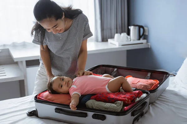 Азиатская мать улыбается держа милого ребенка лежащего в открытом чемодане заполненном — стоковое фото
