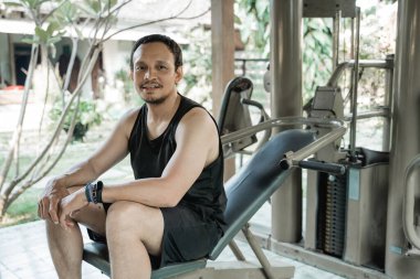 Gülümseyen Asyalı genç adam fitness makinesinde oturuyor.